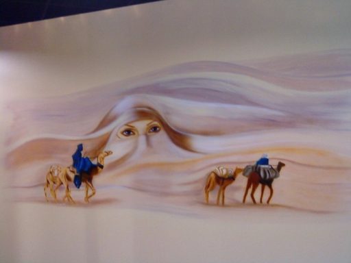 Künstlerische Darstellung einer Kamelkarawane in der Wüste , mit einem Frauengesicht welches in einer Sanddüne erscheint