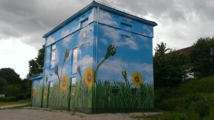 Stromhaus mit Wiese, Sonnenblumen und "gewachsenen" Steckern zum Thema grüner Strom